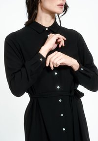 Mustame varrukatega kleit krae, nööpidega ees ja vöökoha lipsuga. Omab säravaid nööpe ja pehmet, sujuvat tekstuuri.