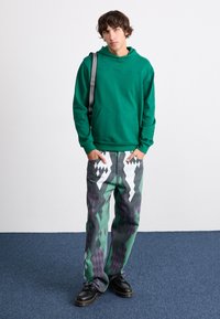 Green cotton hoodie with a front pocket, paired with textured jeans featuring a geometric pattern in grey, white, and purple. Black shoes.