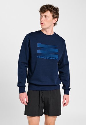 Jonge man draagt een marineblauwe trui met blauwe 'PERFORMANCE' tekst en zwarte korte broek, kijkt naar links tegen een effen achtergrond.