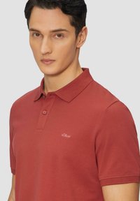 Polo rouge en tissu texturé avec un col, présentant un logo discret sur le côté gauche et deux boutons au niveau du col.