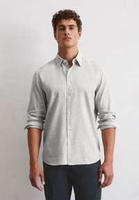 Chemise grise clair à boutons, en tissu texturé, avec un col et des manches retroussées, assortie à un pantalon foncé.