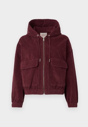 Veste en velours côtelé bordeaux avec capuche, fermeture éclair frontale, deux grandes poches et poignets élastiques. Présente une texture à côtes verticales.