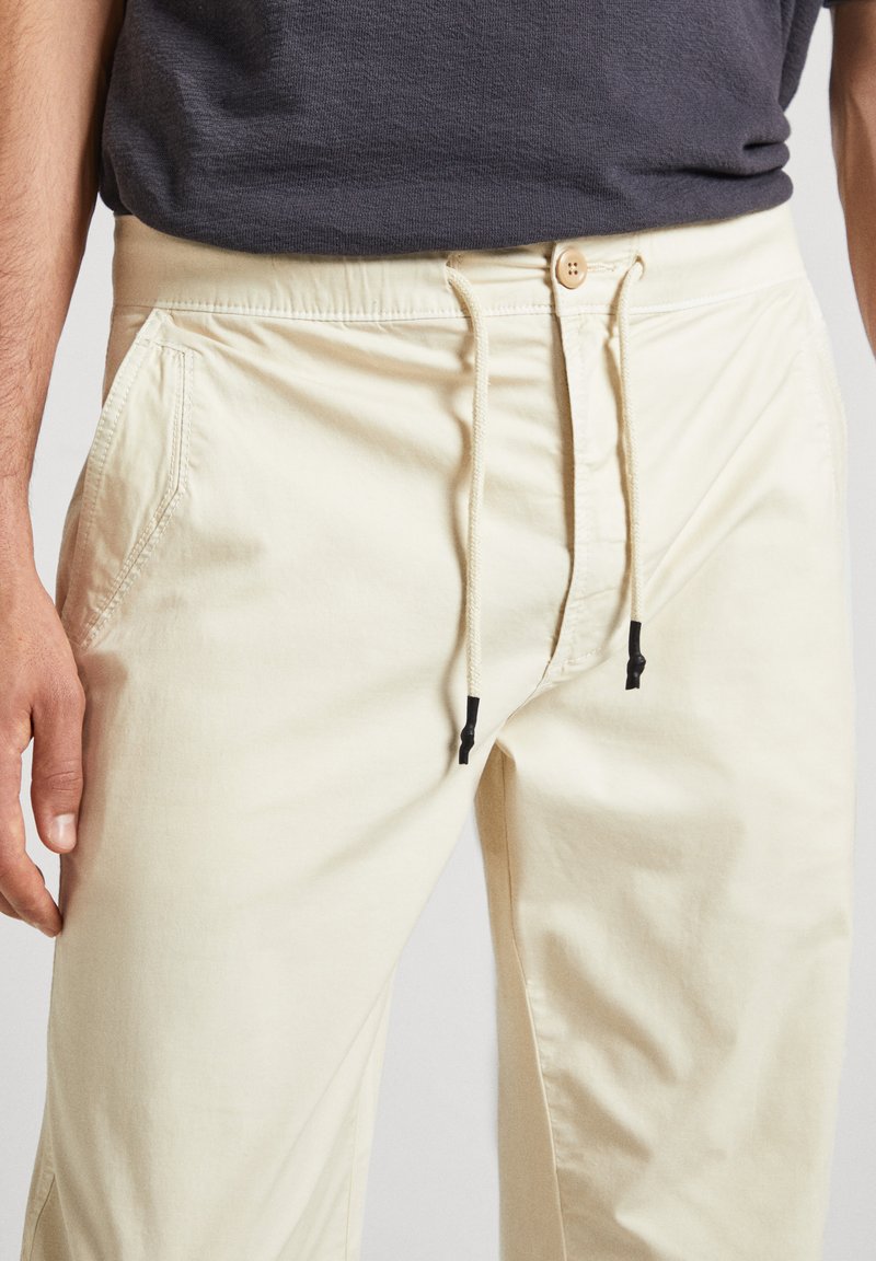 Pantalones color crema hechos de una tela ligera con una cintura ajustable mediante cordón, que cuentan con bolsillos laterales y toggles negros contrastantes.