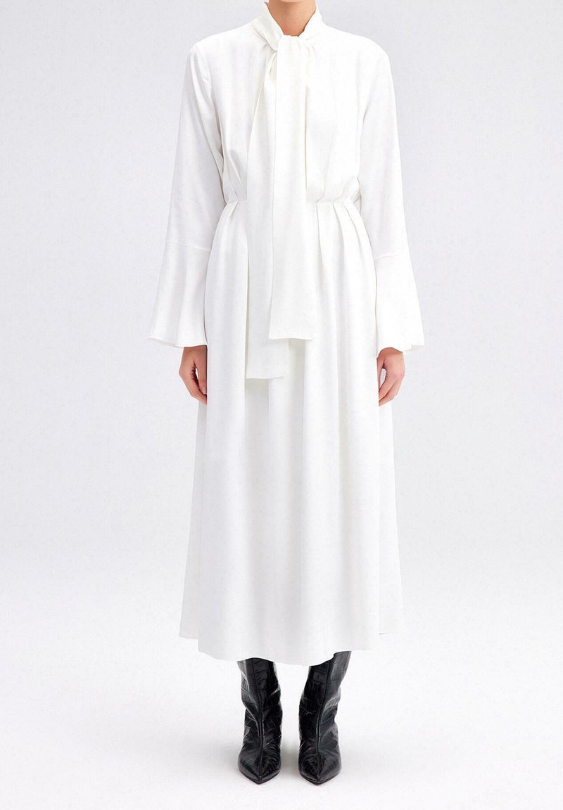 Femme portant une longue robe blanche avec un col noué et des manches évasées, associée à des bottes noires à bout pointu sur un fond clair.