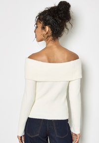 Sweter typu off-the-shoulder, ribbowany, wykonany z miękkiego, jasnobeżowego materiału, z długimi rękawami i marszczonym dekoltem, w połączeniu z ciemnoniebieskimi dżinsami.