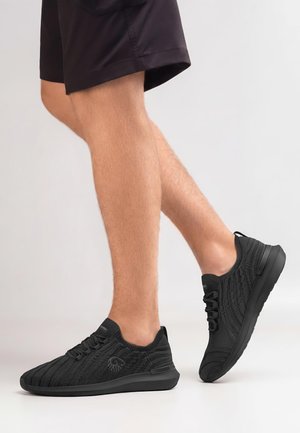 Beine mit schwarzen Stricksneakers und schwarzen Shorts, ein Fuß flach auf dem Boden, der andere mit angehobenem Absatz, hellgrauer Hintergrund.