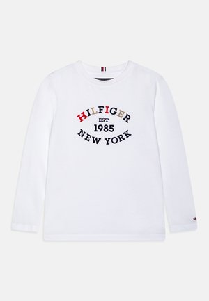 Camisa blanca de manga larga con "HILFIGER EST. 1985 NEW YORK" impreso en letras de colores y negras en el frente, pequeño logo en el puño de la manga izquierda.