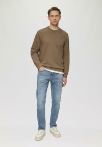 Bruine sweatshirt met ronde hals, gecombineerd met lichtblauwe jeans. Schoenen zijn witte sneakers met beige accenten. Model staat tegen een effen achtergrond.