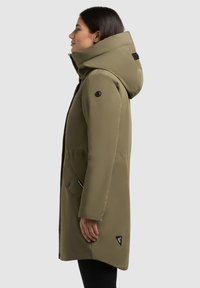 Oliwkowa parka z wysokim kołnierzem i dużym kapturem, z dwoma przednimi kieszeniami, czarnymi guzikami akcentującymi oraz trójkątnym logo na dole.