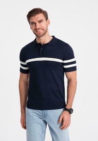 Mörkblå polo skjorta med vita ränder på korta ärmar, tillverkad av stickat tyg. Har en krage och tre-knappsförsedd placket. Avslappnad passform.