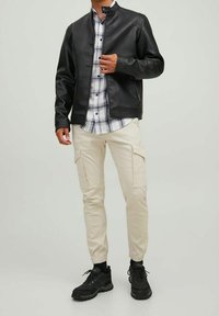 Giacca di pelle nera con colletto rialzato, camicia a quadri, pantaloni cargo beige con tasche e sneakers nere. Outfit moderno e casual.