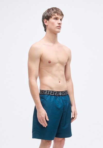 Τιρκουάζ μαγιό σορτς με μαύρη μέση με γεωμετρικές λεπτομέρειες, με άνετη εφαρμογή και λεία υφή υφάσματος, τοποθετημένο σε μοντέλο.