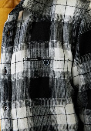 Chemise en flanelle à carreaux noirs et blancs avec une poche poitrine boutonnée portant l'étiquette "Garcia", portée sur un vêtement en tricot jaune moutarde en dessous.