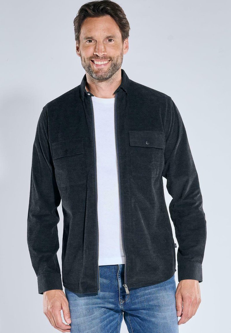 Dunkelgraues Cordhemd mit einem Reißverschluss an der Vorderseite, zwei Brusttaschen und einem kragenlosen Ausschnitt, getragen über einem weißen T-Shirt und blauen Jeans.
