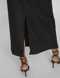 Abito in satin nero di lunghezza media, con uno spacco laterale, abbinato a sandali con tacco alto e cinturini neri. Texture liscia.