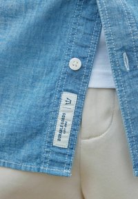 Chemise en denim bleu clair avec un bouton blanc, une étiquette latérale portant la mention "DURABLE GOODS" et un tissu de couleur crème en dessous. Texture douce, détails cousus.