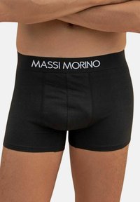 Uomo che indossa boxer neri con un elastico "MASSI MORINO", braccia incrociate sul torso nudo.
