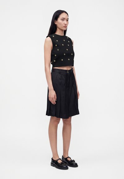 Γυναίκα που φοράει μαύρο αμάνικο cropped top με χρυσά μεταλλικά κουμπώματα, μαύρη πλισέ φούστα μέχρι το γόνατο και μαύρα παπούτσια με αγκράφες σε λευκό φόντο.