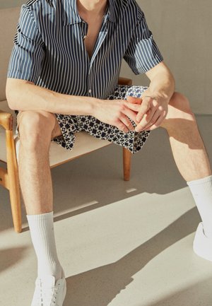 Muž sedící na dřevěné židli, oblečený v modré pruhované košili s krátkými rukávy, vzorovaných šortkách, bílých ponožkách a bílých teniskách na slunečním světle.