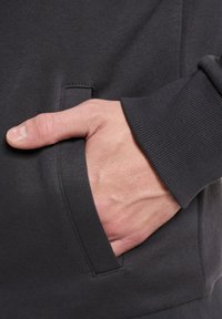 Felpa grigio scuro con tasca laterale e polsini a coste. Ingrandimento di una mano appoggiata nella tasca, che mette in evidenza la texture e i dettagli del tessuto.