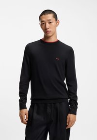 Maglione nero con colletto rotondo, maniche lunghe e accentuazione rossa sul colletto. Presenta un piccolo logo "HUGO" rosso sul petto, realizzato in un materiale liscio.
