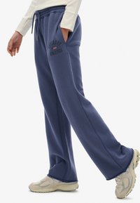 Persona vistiendo pantalones deportivos Superdry Athletics azules con cintura elástica y zapatillas beige, caminando frente a un fondo blanco liso.