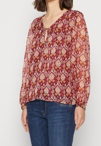 Donna che indossa una blusa trasparente con maniche lunghe, con un motivo geometrico rosso, rosa e senape annodata al collo, abbinata a jeans di denim blu.
