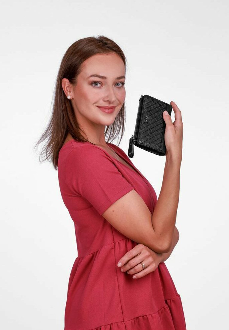 Černá prošívaná peněženka držena v levé ruce, s uzávěrem na zip a malým střapcem. Modelka má na sobě růžové šaty s krátkým rukávem.