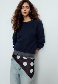 Maglione a maglia testurizzata blu navy abbinato a una camicia a righe e a un tessuto triangolare scuro con pois azzurri, indossato sopra jeans in denim.