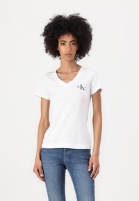 White short-sleeve V-neck T-shirt with a small black logo on the left side, made from soft cotton material. Worn with blue jeans.