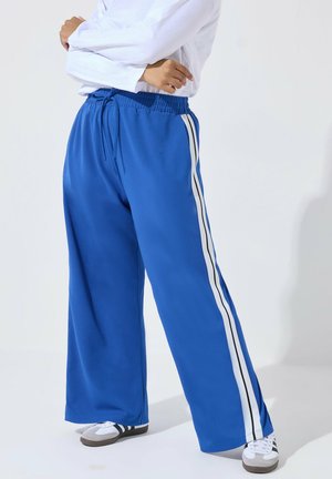 Persona che indossa pantaloni blu a gamba larga con strisce laterali bianche e nere, maglia bianca a maniche lunghe e sneakers bianche, in piedi con le braccia incrociate.
