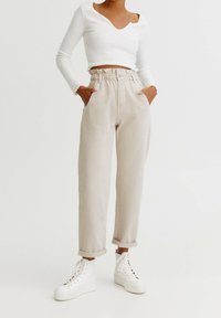 Pantalones beige de cintura alta con cinturilla elástica fruncida, bolsillos laterales y dobladillos acampanados, combinados con una camiseta corta de manga larga blanca y zapatillas blancas.