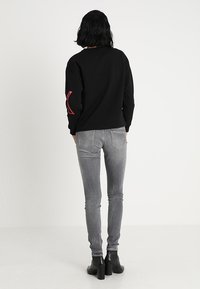 Černá mikina s červenými akcenty na rukávech, kombinovaná se šedými úzkými džínami a černými kotníkovými botami, pohled zezadu na bílém pozadí.