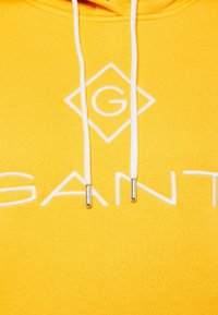 Sudadera amarilla con un logo bordado en blanco, que presenta una forma de diamante y la palabra "GANT". Cuerdas blancas con puntas metálicas.