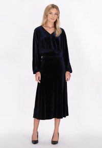 Lange marineblauwe fluwelen jurk met een V-hals, lange mouwen en een getailleerde taille, uitlopende in een losse rok, gecombineerd met zwarte hakken.