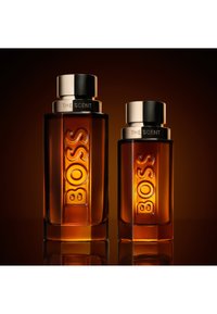 Deux flacons de parfum en verre ambré avec des bouchons argentés, étiquetés "BOSS THE SCENT", présentés sur un fond dégradé marron foncé.