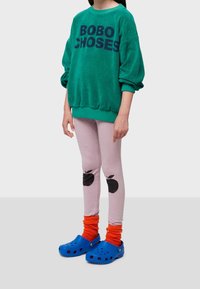 Niño con una camiseta de manga larga verde con el texto "BOBO CHOSES", mallas rosa claro con estampado de manzanas negras, calcetines naranjas y zuecos azules.