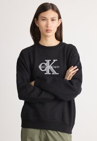 Čierna bavlnená mikina s rebrovaným golierom a manžetami, s výrazným šedým logom "CK" a textom "Calvin Klein" v strede.