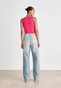 Γυναίκα με τα μαλλιά σε σινιόν, φορώντας αμάνικο ροζ τοπ και φαρδιά ξεθωριασμένα μπλε τζιν, να στέκεται με την πλάτη γυρισμένη σε έναν απλό τοίχο.