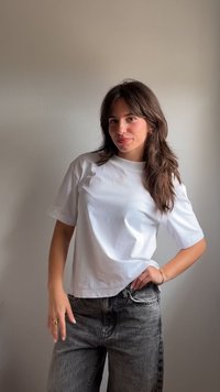 Νεαρή γυναίκα με μακριά σκούρα μαλλιά, φοράει ένα λευκό t-shirt και γκρι τζιν και στέκεται μπροστά σε έναν απλό ανοιχτό γκρι τοίχο, χαμογελώντας ελαφρώς.