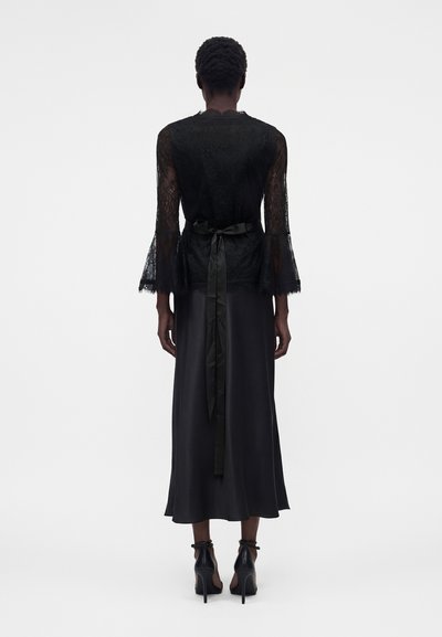 Άτομο που φοράει μαύρο δαντελένιο τοπ με μανίκια καμπάνα και σατέν κορδέλα δεμένη στη μέση, συνδυασμένο με μαύρη midi φούστα και ψηλά τακούνια.