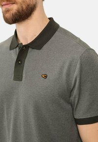 Graues Poloshirt mit einem dunkelgrünen Kragen, einer Knopfleiste mit drei Knöpfen und einem kleinen gelben Logo, das auf der Brust gestickt ist. Strukturierter Stoff sichtbar.