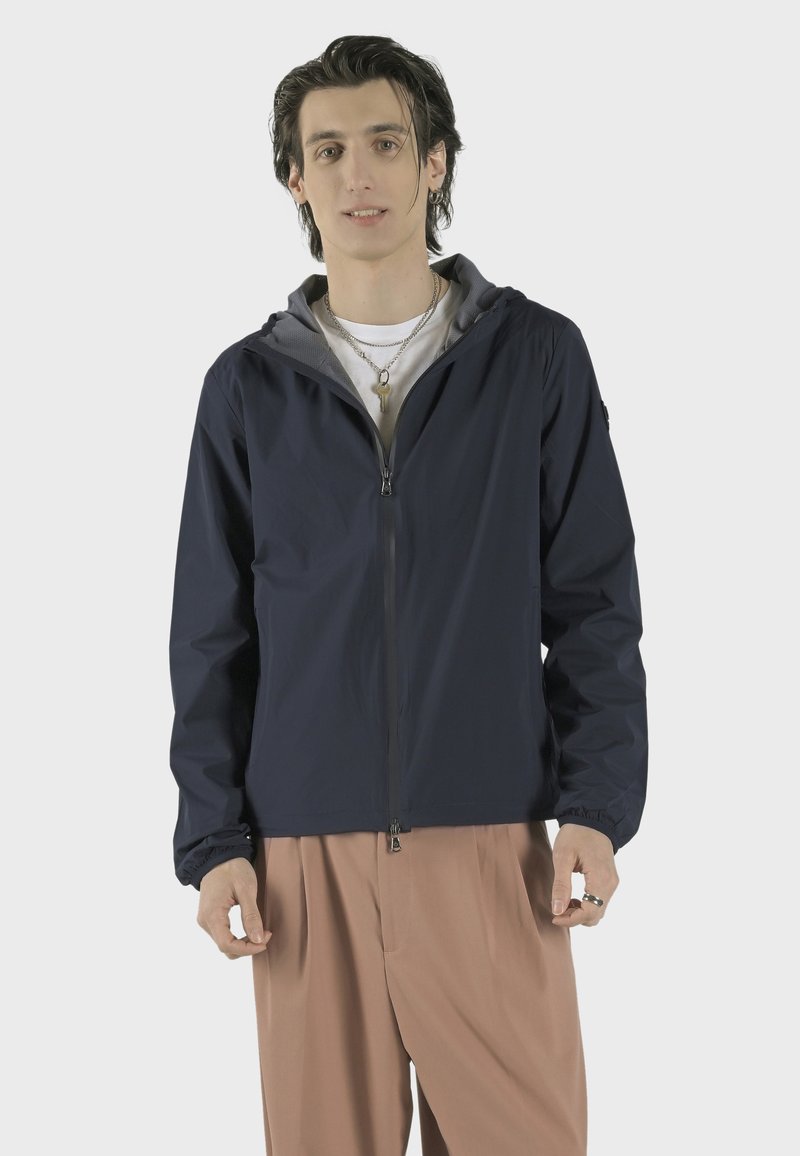 Námořnická lehká bunda se šedou podšívkou kapuce, předním zipem, elastickými manžetami a volným střihem; nošena přes bílé tričko a béžové kalhoty.