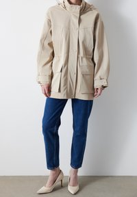 Beżowa wodoodporna kurtka z kapturem, z przednimi kieszeniami i zatrzaskami. Noszona na niebieskich dżinsach i jasnych szpilkach.