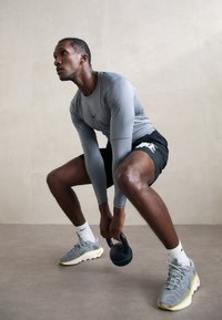 Vyras pilku ilgomis rankovėmis marškiniais ir juodais šortais atsisėdęs laiko juodą kettlebell'ą, ruošiasi treniruotei šviesiame grindų paviršiuje.