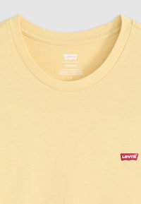 Camiseta de algodón amarilla claro con cuello redondo y pequeño logo rojo de Levi's cosido en el lado izquierdo. Textura suave con un diseño sencillo.