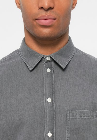 Chemise grise à manches longues en tissu doux, dotée d'un col pointu, de boutons blancs et d'une poche poitrine. Design simple avec une finition texturée.