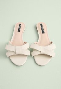 Beige skidesandaler med en glat læderoverflade, der har en fremtrædende bueaccent på forsiden. Blød indersål, minimalisk design.