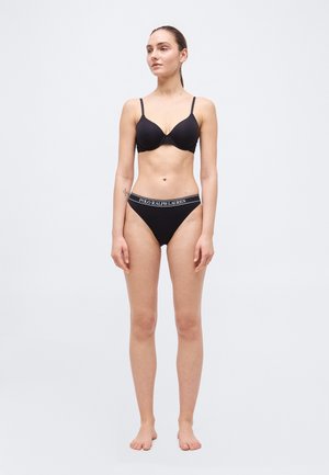 Mujer de pie descalza, vestida con sujetador negro y ropa interior negra con cinturilla que lleva la marca "Polo Ralph Lauren", contra un fondo claro y liso.