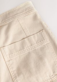 Tissu en denim beige présentant une poche arrière avec double coutures et un bord net, mettant en valeur une surface lisse et texturée.